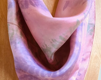 Könnte beinhalten: Ein rosa Seidentuch mit einem hellvioletten und grünen Blumenmuster. Das Tuch ist gefaltet und auf einer Holzoberfläche ausgestellt.