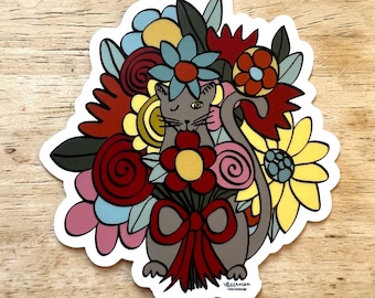 Könnte beinhalten: Eine graue Katze mit einem zwinkernden Auge liegt in einem Strauß bunter Blumen. Die Blumen sind rot, gelb, rosa und blau. Die Katze trägt eine rote Schleife. Der Sticker hat den Text "victoria:design" unten.