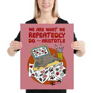 May include: A pink poster with a quote from Aristotle: "We are what we repeatedly do." The quote is illustrated with a gray cat sitting on a laptop, surrounded by paintbrushes, paint, and paper with hearts and paw prints.