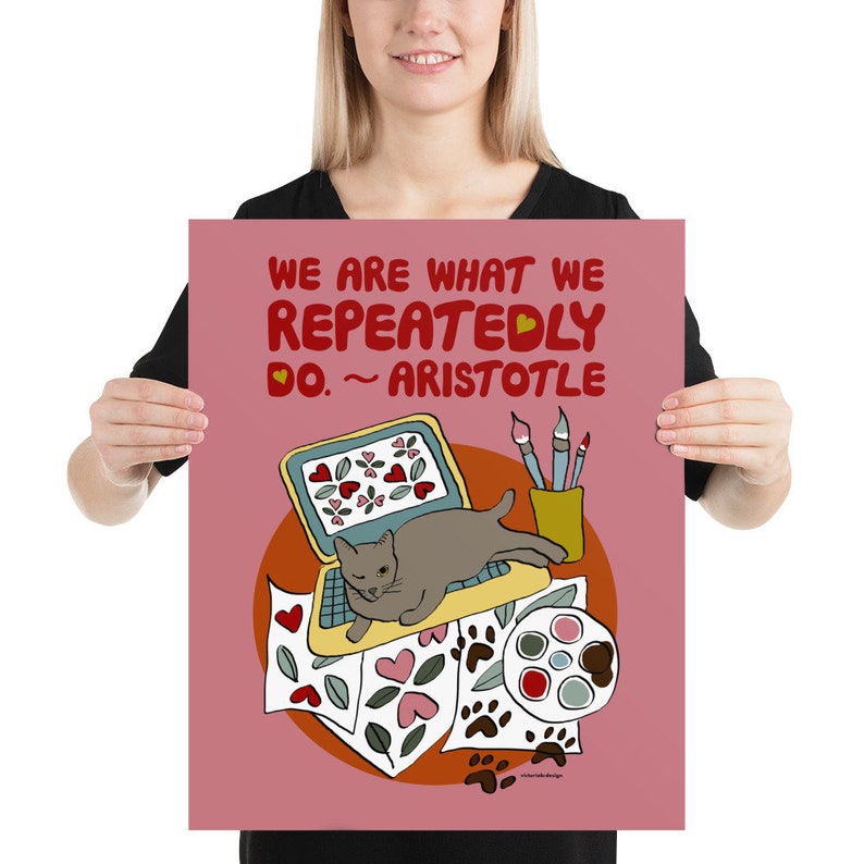 May include: A pink poster with a quote from Aristotle: "We are what we repeatedly do." The quote is illustrated with a gray cat sitting on a laptop, surrounded by paintbrushes, paint, and paper with hearts and paw prints.