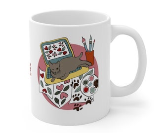 Könnte beinhalten: Weiße Keramiktasse mit einer bunten Illustration einer Katze auf einem Laptop, Pinseln, einer Farbpalette und Pfotenabdrücken. Die Illustration ist von einem rosa Kreis umgeben.