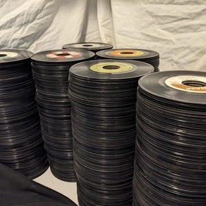 May include: A stack of vintage vinyl records in various colors, including black, white, red, and yellow. The records are stacked high, creating a tower of music.