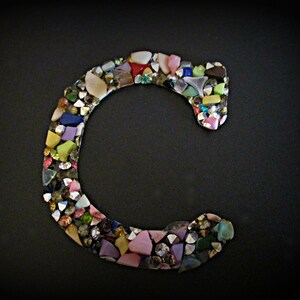 May include: A colorful mosaic letter C made from small pieces of glass and other materials. The letter is on a black background.
