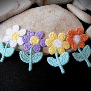 May include: Four embroidered flower patches in white, purple, yellow, and orange, each with a yellow or white center. Light blue leaves and stems accompany each flower. The patches are arranged on a dark surface.