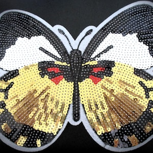 May include: A sequined butterfly applique with a black, white, gold, and red color scheme. The butterfly has a gray border and is likely for decorative purposes, such as embellishing clothing or accessories.