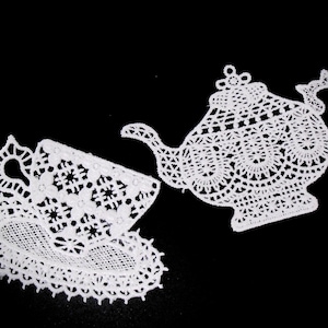 May include: Two white lace appliques featuring intricate needlework designs. One piece is shaped like a teapot with a lid and handle, while the other is shaped like a teacup resting on a saucer, both displaying detailed openwork patterns.