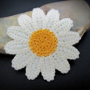 May include: A handmade crochet daisy with white petals and a yellow center. The flower is placed on a gray stone against a dark background. This floral applique is suitable for various craft projects.