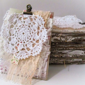 May include: A stack of handmade paper pages with lace and crocheted doilies, bound together with a metal clip. The pages are decorated with various textures and patterns, creating a vintage and romantic aesthetic.