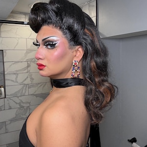 Op de afbeelding: Een persoon met opvallende make-up, waaronder gedurfde oogschaduw en rode lippenstift, draagt grote, kleurrijke oorbellen. Het haar is gestyled in een volumineuze updo met donkere en kastanjebruine highlights. Een zwarte choker en een zwarte jurk maken de look compleet.