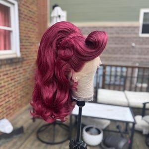 Może przedstawiać: Burgundowa peruka w stylu vintage, z falami i lokami. Peruka jest umieszczona na głowie manekina, prezentując skomplikowany design i kolor. Włosy są ułożone z przedziałkiem z boku i zaplecionym detalem.