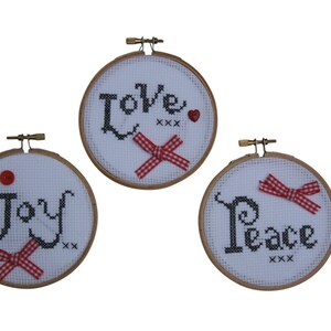 Puede incluir: Tres aros de bordado redondos de madera con tela blanca estirada sobre ellos. Cada aro tiene una palabra diferente bordada en hilo negro: "Love", "Joy" y "Peace". Cada aro tiene una cinta roja y blanca atada alrededor.
