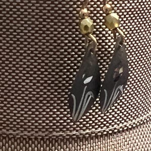 May include: A pair of dark grey teardrop earrings with white painted accents. The earrings hang from gold-coloured hooks and are adorned with small, olive-green beads. The background is a textured brown fabric.
