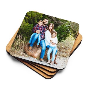 May include: A stack of square coasters with a cork base. The top coaster features a family portrait with a man, woman, and two children. The family is sitting on a rock in a natural outdoor setting. The coasters are square with rounded corners.