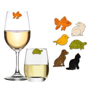 May include: Wine glasses with white wine and wooden animal charms. The charms include a goldfish, rabbit, bird, turtle, dog, and cat. The goldfish and turtle charms are attached to the glasses.