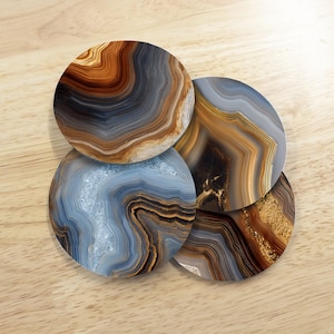 May include: Four round coasters with agate stone designs. The coasters feature swirling patterns in shades of brown, gold, gray, and blue. The coasters are arranged on a light wood surface.