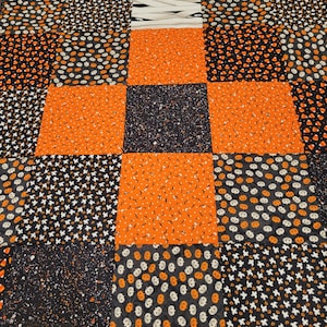 May include: A patchwork quilt with a Halloween theme. The quilt is made of various fabrics with different patterns, including pumpkins, bats, candy corn, and ghosts. The quilt is in shades of black, orange, and white.