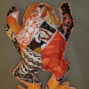 May include: A patchwork frog toy made from colorful fabric scraps. The frog has orange, brown, white, and black fabric with a floral pattern. The frog has large orange eyes.