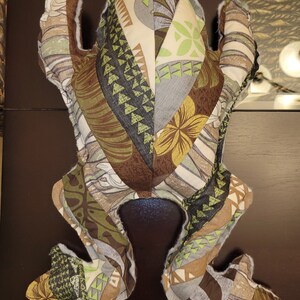 May include: A plush frog toy made of patchwork fabric with a brown, green, and white geometric pattern. The frog is lying on its back with its legs outstretched.