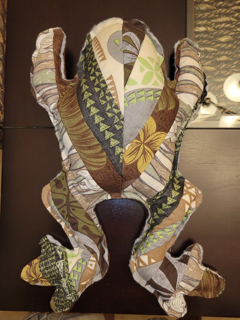 May include: A plush frog toy made of patchwork fabric with a brown, green, and white geometric pattern. The frog is lying on its back with its legs outstretched.