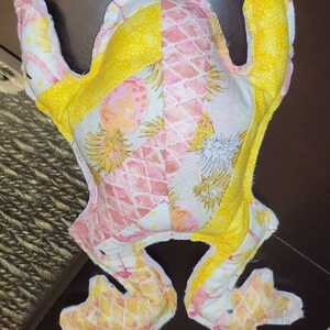 May include: A yellow and pink frog plush toy with a pineapple print. The frog is made of soft fabric and has a quilted design.