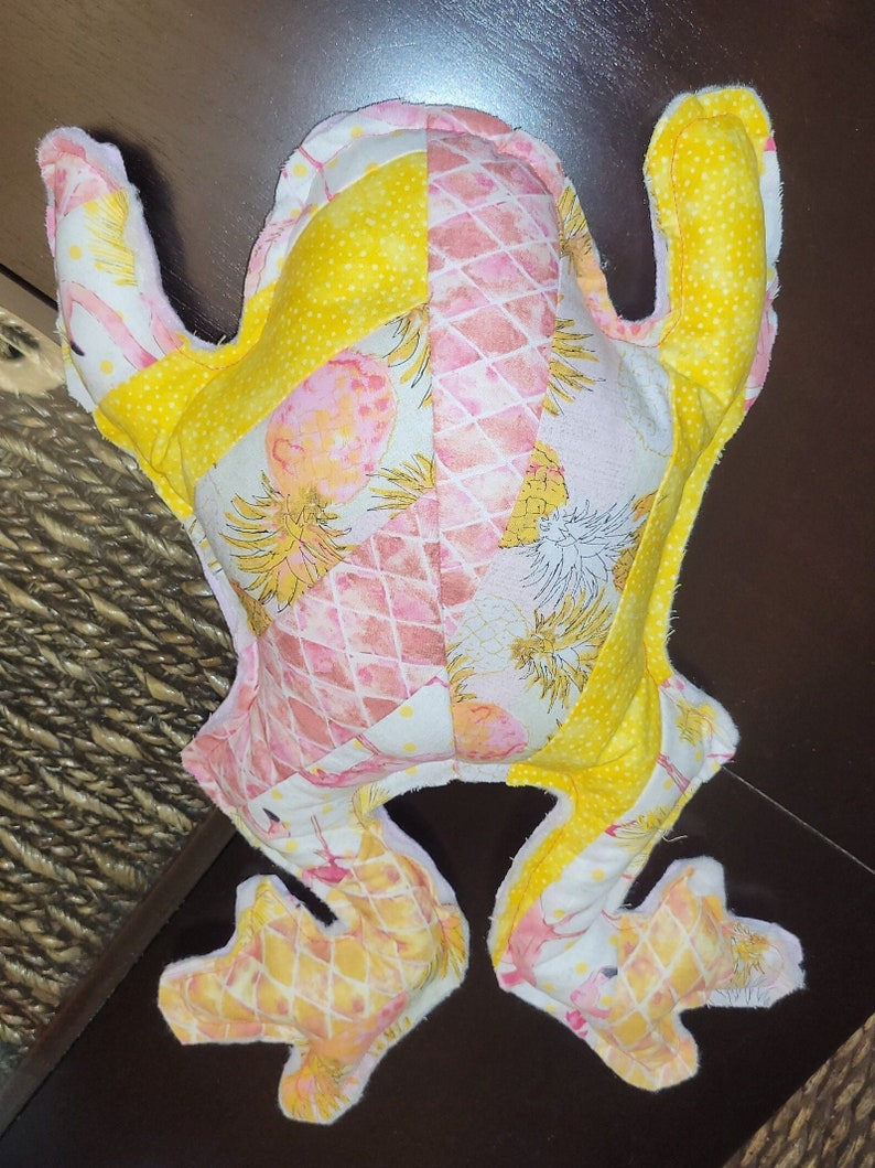 May include: A yellow and pink frog plush toy with a pineapple print. The frog is made of soft fabric and has a quilted design.