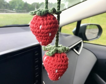 May include: Two red crocheted strawberry car charms with green leaves and white seed details, hanging from a green braided cord. The charms are suspended in front of a car's dashboard and rearview mirror.
