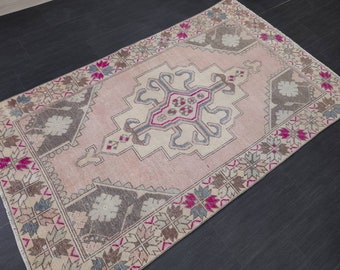 Puede incluir: Alfombra rectangular con un diseño geométrico central en crema, gris y rosa. La alfombra tiene un fondo rosa y un borde con patrones florales y geométricos en tonos marrones, rosas y azules. La alfombra probablemente está hecha de lana o un material similar.