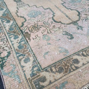 May include: A vintage rug with a faded, distressed appearance. The rug features a central field of pale pink with floral and geometric patterns in shades of green, brown, and cream. A dark green border frames the design.
