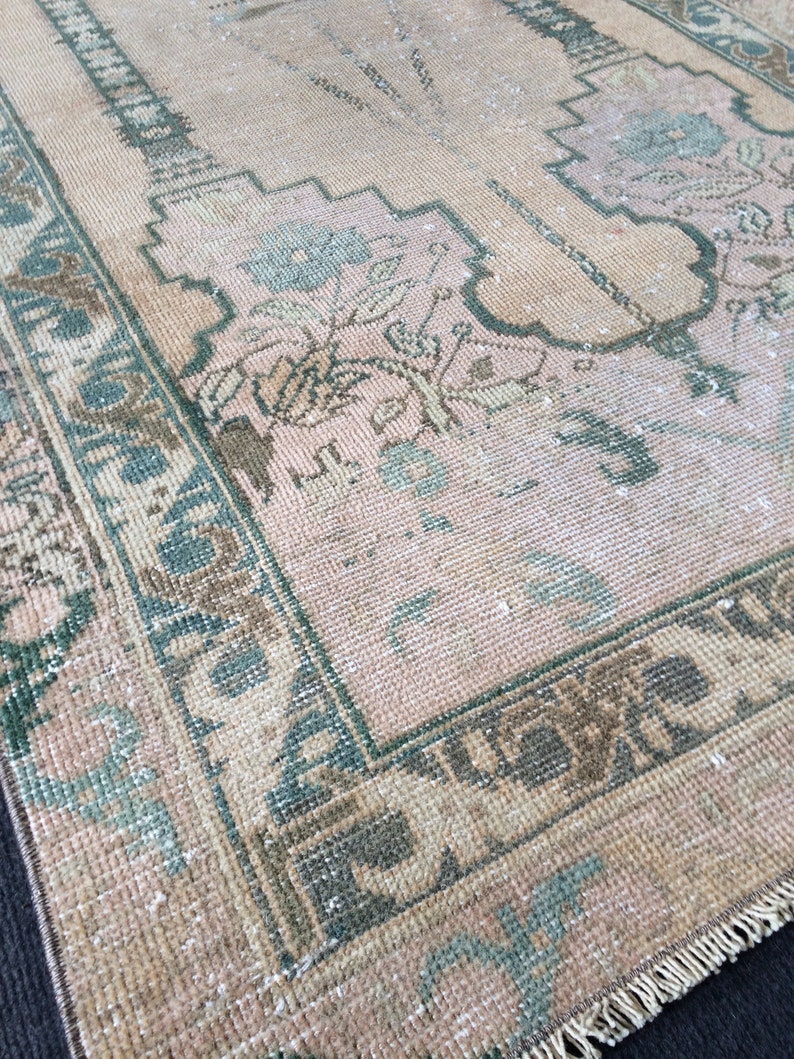 May include: A vintage rug with a faded, distressed appearance. The rug features a central field of pale pink with floral and geometric patterns in shades of green, brown, and cream. A dark green border frames the design.