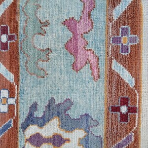 May include: Close-up of a colorful rug with a textured surface. The rug features a light blue background with brown, purple, and pink geometric patterns. The edges have a cream-colored border and fringe.