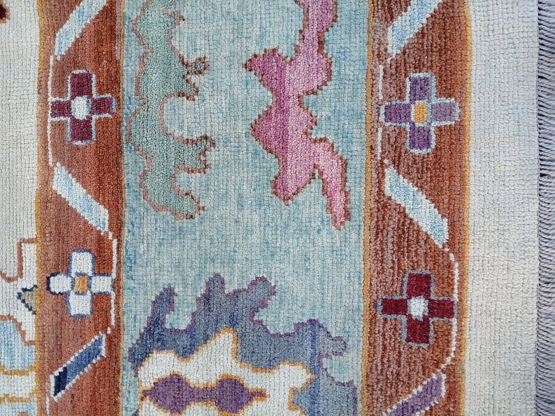May include: Close-up of a colorful rug with a textured surface. The rug features a light blue background with brown, purple, and pink geometric patterns. The edges have a cream-colored border and fringe.