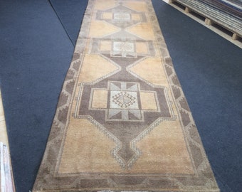 May include: A rectangular, beige and brown patterned runner rug. The rug features geometric designs in shades of brown and beige, with a central medallion and border details. The rug is displayed on a dark blue surface.