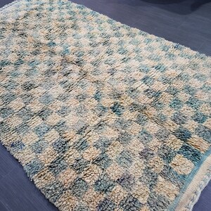 Op de afbeelding: Handgemaakt tapijt met een ruitpatroon in cr&egrave;me, blauw en beige tinten. Het tapijt heeft een getextureerd oppervlak en een gefranjerde rand. Het ontwerp heeft een herhalend vierkant patroon.