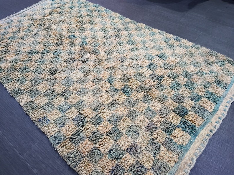 Op de afbeelding: Handgemaakt tapijt met een ruitpatroon in cr&egrave;me, blauw en beige tinten. Het tapijt heeft een getextureerd oppervlak en een gefranjerde rand. Het ontwerp heeft een herhalend vierkant patroon.