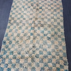 Op de afbeelding: Een rechthoekig vloerkleed met een geblokt patroon in blauw- en cr&egrave;metinten. Het vloerkleed heeft een harige textuur en een franje langs de onderrand. Het is een decoratief woondecoratie-item.