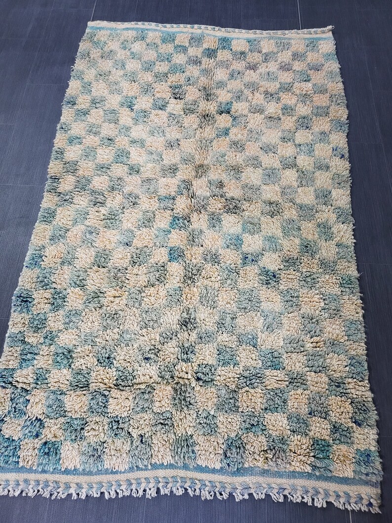 Op de afbeelding: Een rechthoekig vloerkleed met een geblokt patroon in blauw- en cr&egrave;metinten. Het vloerkleed heeft een harige textuur en een franje langs de onderrand. Het is een decoratief woondecoratie-item.