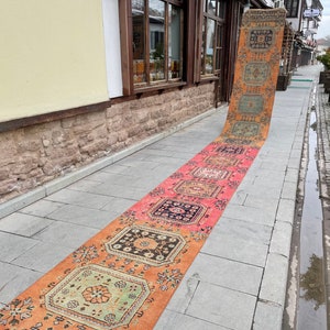May include: A long, rectangular, vintage rug with a repeating geometric pattern in shades of orange, pink, and blue. The rug is displayed outdoors on a stone walkway, showcasing its intricate design and warm color palette.