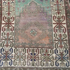May include: A small, rectangular, vintage rug with a central diamond design in shades of purple, orange, and teal. The rug features intricate geometric patterns in red, blue, and green, framed by a border of repeating motifs. The rug has a distressed appearance.
