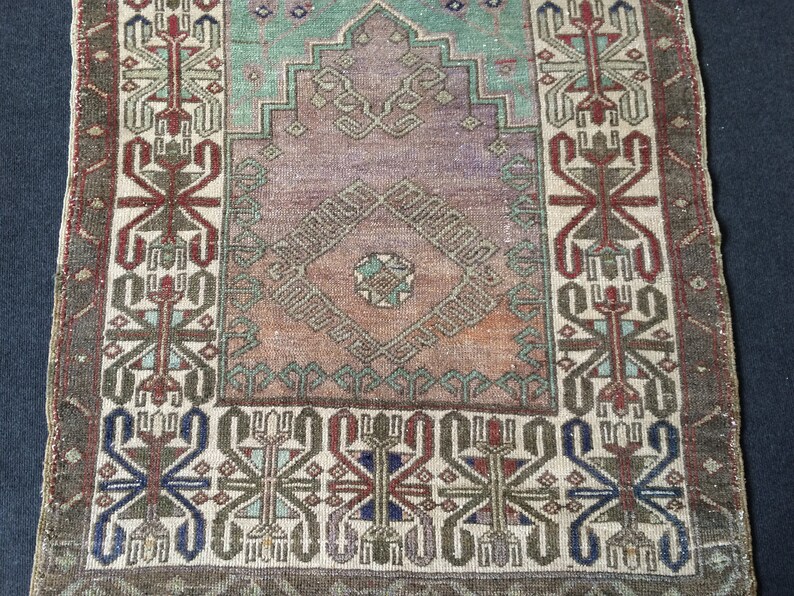 May include: A small, rectangular, vintage rug with a central diamond design in shades of purple, orange, and teal. The rug features intricate geometric patterns in red, blue, and green, framed by a border of repeating motifs. The rug has a distressed appearance.