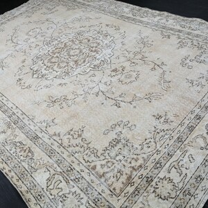 Puede incluir: Una alfombra rectangular grande con un intrincado diseño floral en tonos beige y marrón. La alfombra presenta un diseño de medallón central y un borde detallado, creando una estética vintage. La alfombra está colocada sobre una superficie oscura.