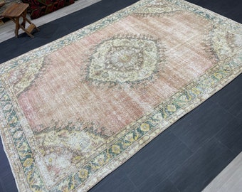 May include: A rectangular, distressed rug with a central medallion design. The rug features a faded color palette of pink, cream, and green, with intricate floral patterns along the border and within the central design. The rug's dimensions are approximately 6 x 9 feet.