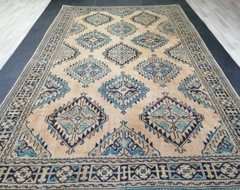 May include: Large beige rug with a repeating diamond pattern in shades of blue and navy. The rug has a detailed border with geometric designs. The overall design is symmetrical and the rug appears to be made of a woven material.