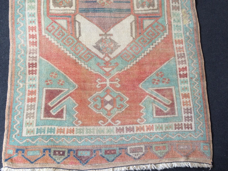 Puede incluir: Una alfombra rectangular de estilo vintage con un dise&ntilde;o central en tonos melocot&oacute;n, turquesa y burdeos. La alfombra presenta patrones geom&eacute;tricos y un borde con flecos. El dise&ntilde;o general es sim&eacute;trico, con una apariencia desgastada.