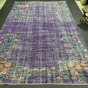 May include: A large, rectangular purple rug with a distressed, vintage appearance. Floral patterns in shades of green, yellow, and pink adorn the edges. The rug's dimensions are not visible in the image.