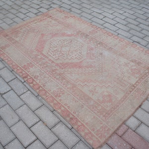 May include: A rectangular, distressed rug with a central geometric design and a border. The rug features a faded, vintage aesthetic with a color palette of beige and muted red. The rug is placed on a brick surface.