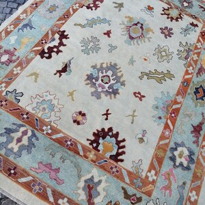 May include: A rectangular area rug with a cream-colored background and a detailed border. The border features a light blue and rust-colored design. The rug has floral and abstract patterns in shades of burgundy, blue, and yellow.
