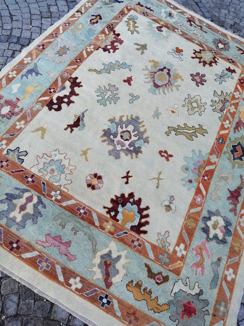 May include: A rectangular area rug with a cream-colored background and a detailed border. The border features a light blue and rust-colored design. The rug has floral and abstract patterns in shades of burgundy, blue, and yellow.