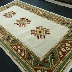 Puede incluir: Alfombra rectangular crema con un borde detallado en tonos verdes, marrones y blancos. La alfombra presenta tres diseños florales centrales en marrón, rojo y amarillo. Las dimensiones de la alfombra no son visibles.