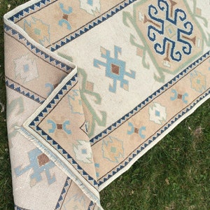 May include: A beige and cream-colored runner rug with a geometric pattern. The rug features a central cream field with a repeating pattern of blue, brown, and green motifs. The border is beige with a dark blue zigzag design.