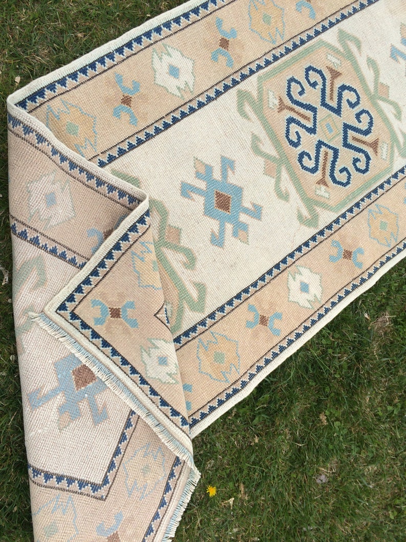 May include: A beige and cream-colored runner rug with a geometric pattern. The rug features a central cream field with a repeating pattern of blue, brown, and green motifs. The border is beige with a dark blue zigzag design.